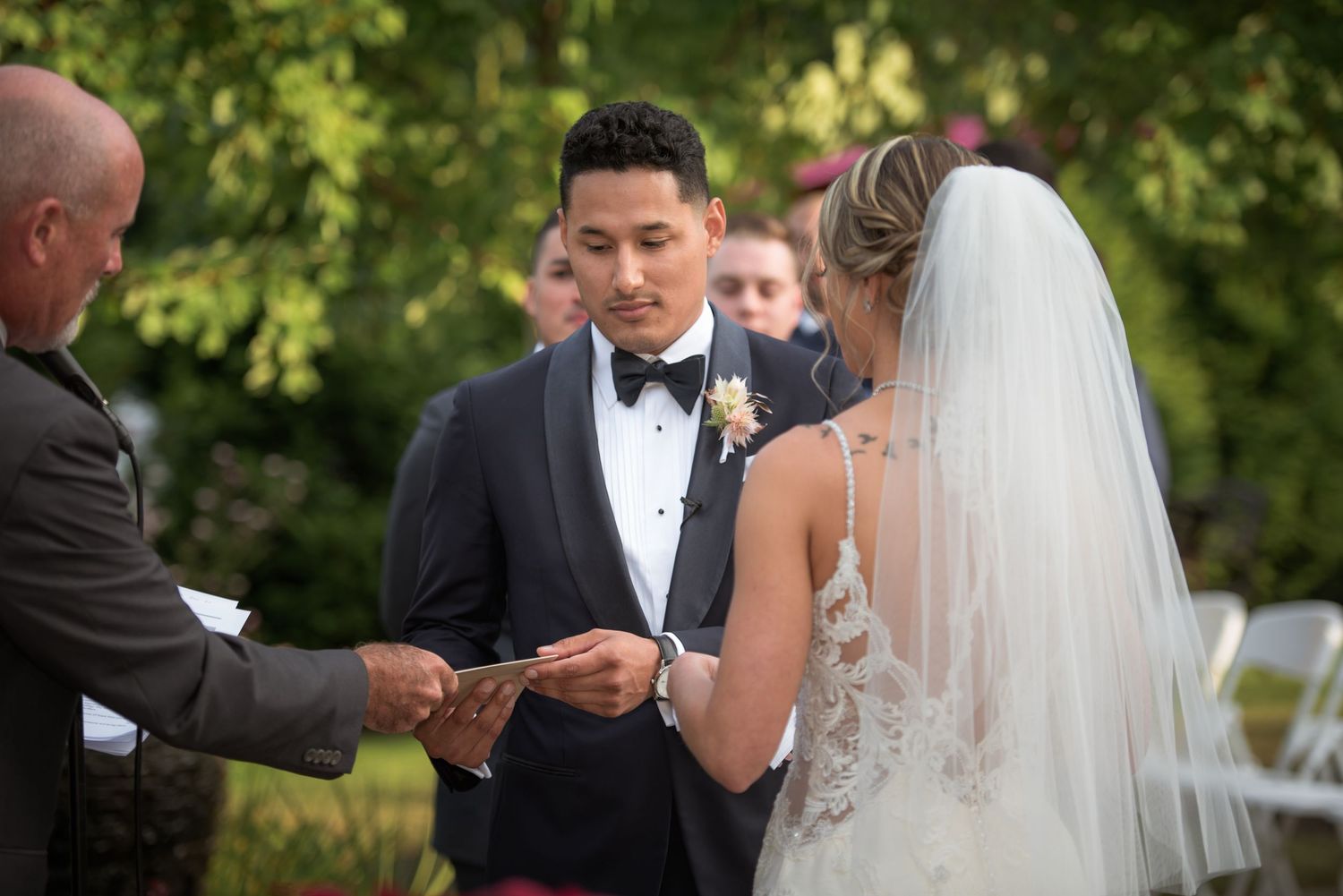 Groom in navy tuxedo receives wedding ring from officiant during outdoor ceremony while bride in lace gown watches.