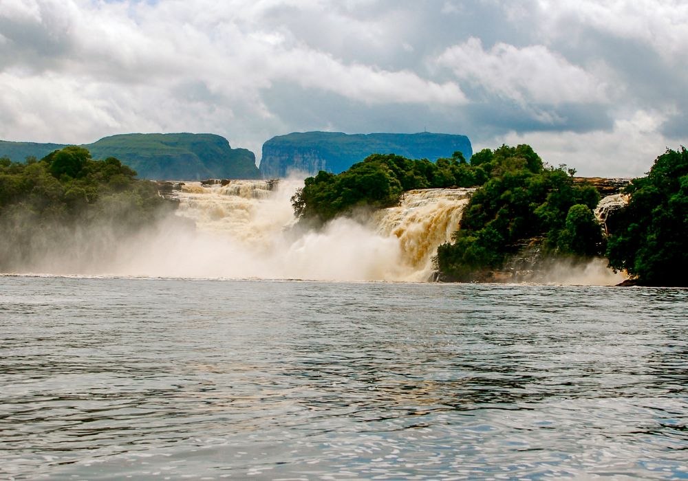 Wide cascading waterfalls thunder over cliffs in a tropical river landscape with mountains in background.