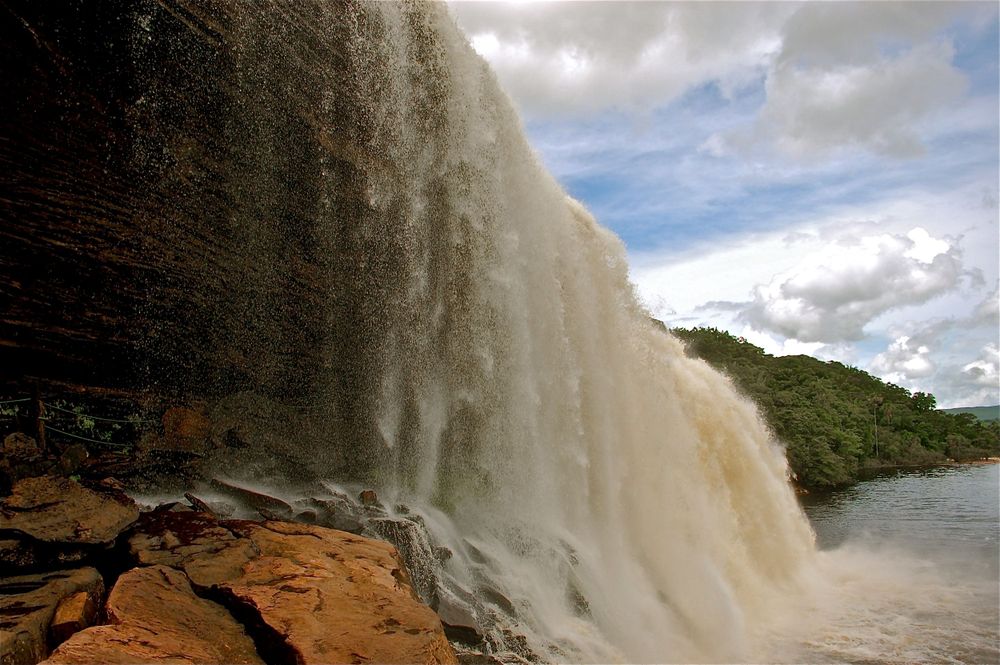 Powerful waterfall cascades over rocky cliffs into a river with green hills visible in the background.
