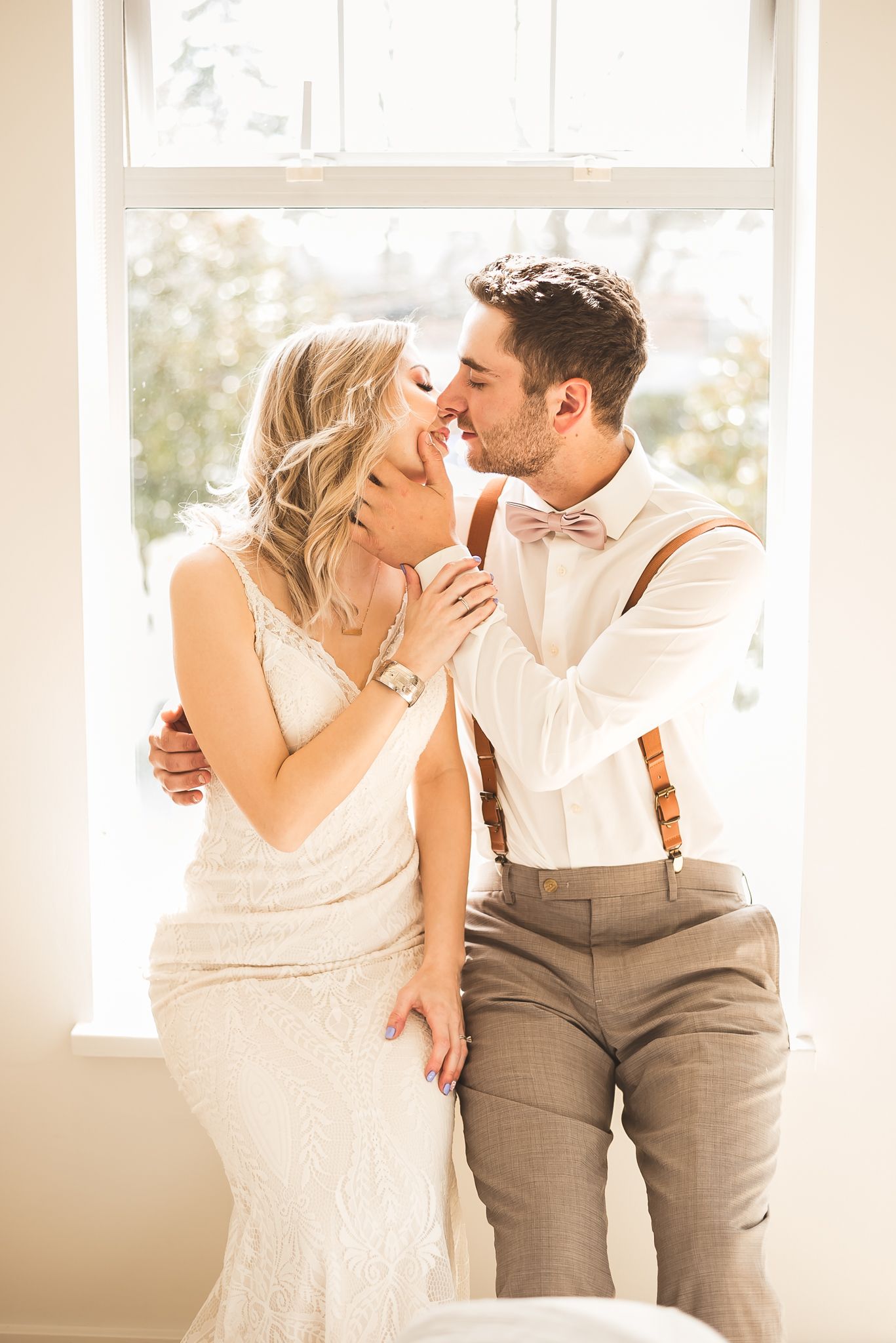 Sitting in front of a window filled with light, a bride and groom share an almost kiss as they celebrate their wedding day.