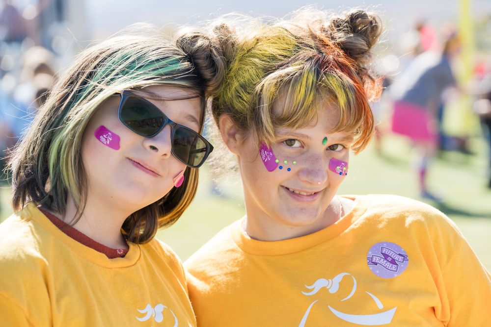 Two participants with colorful face paint and hair celebrate at Girls on the Run event wearing matching yellow shirts.