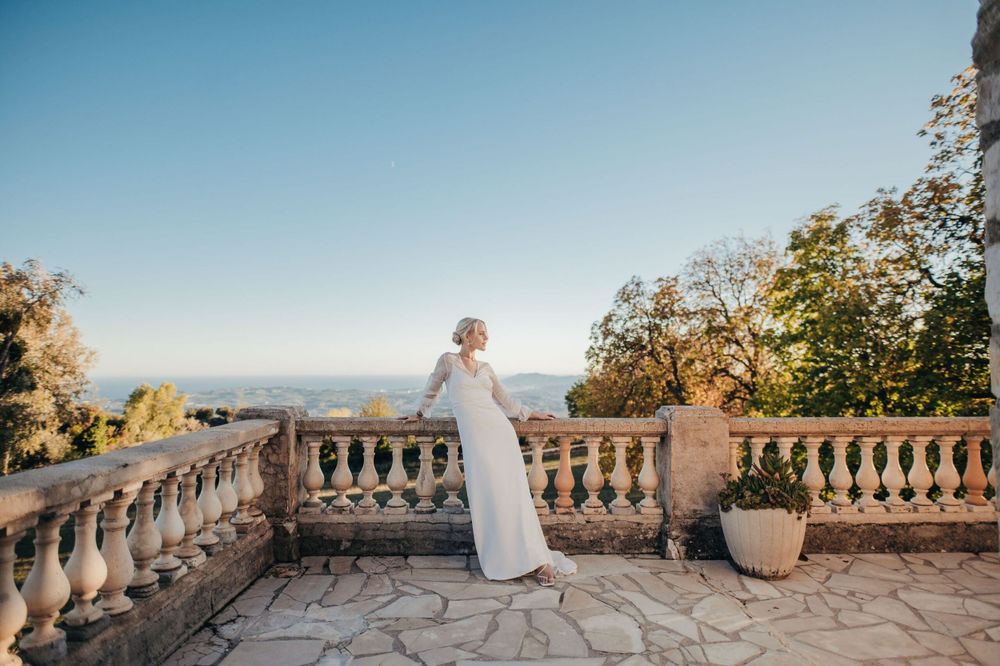Figure in an elegant white dress stands on a stone balcony overlooking a scenic landscape at sunset.