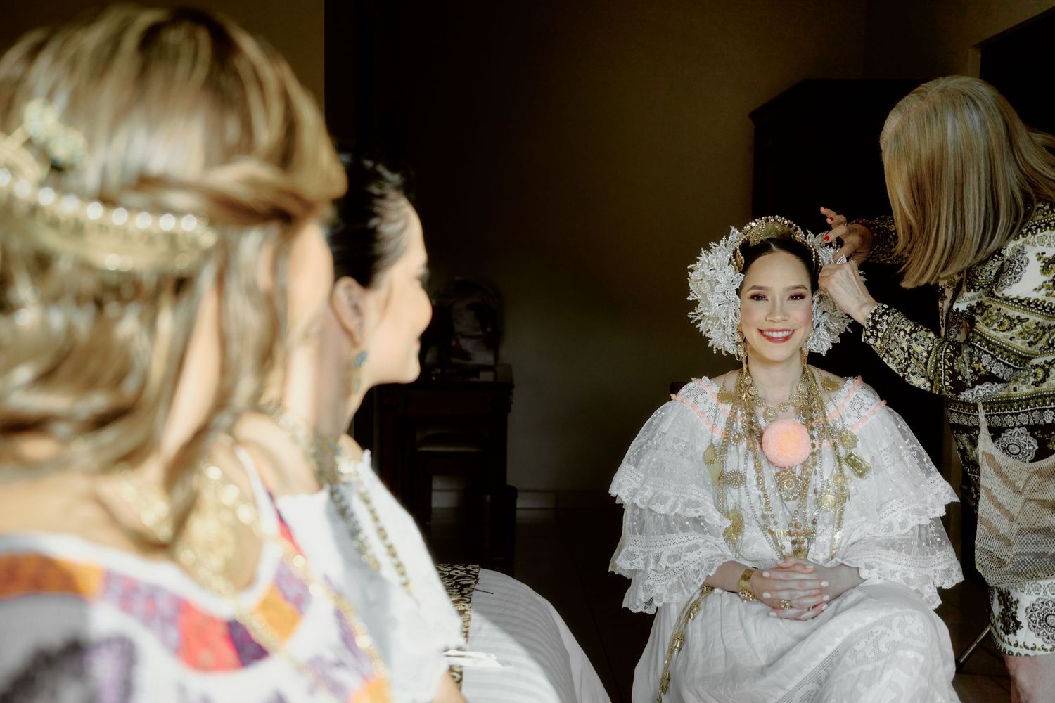 Traditional bride wearing ornate lace dress and headdress surrounded by attendants during ceremony preparation.