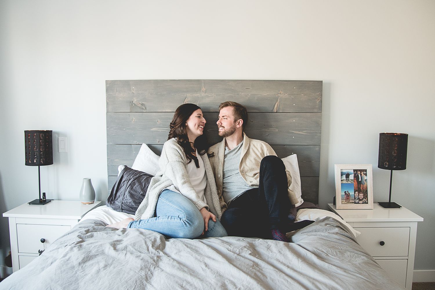 A couple sits intimately on a gray-headboard bed with white bedding and matching nightstands.