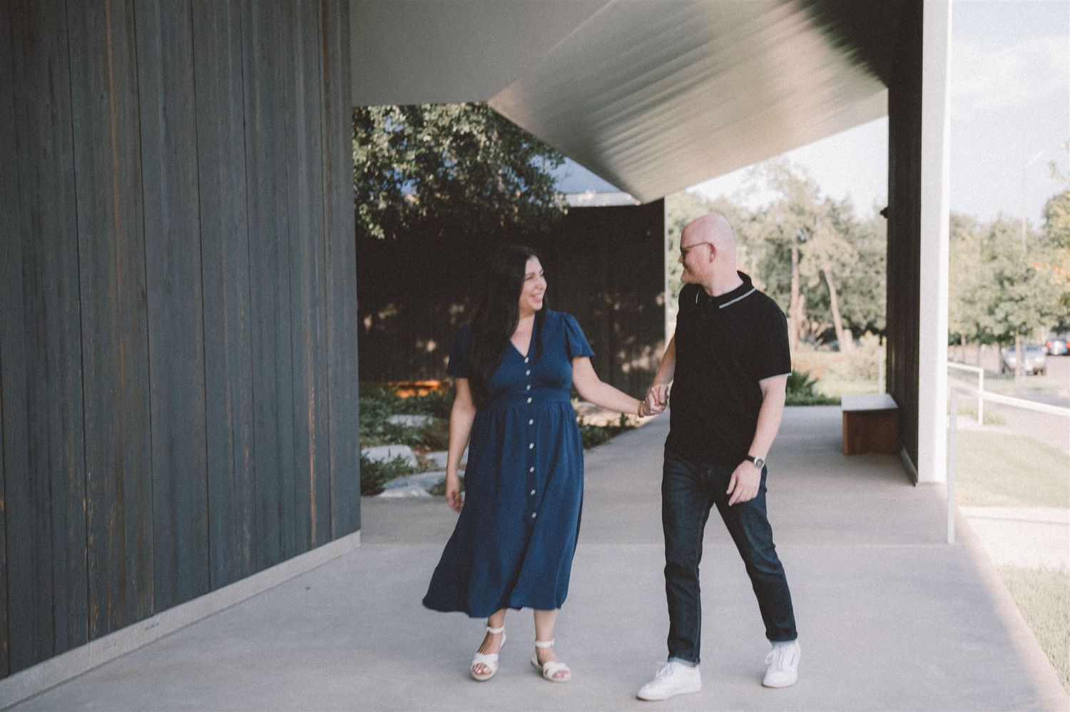 A couple in contrasting outfits walks together under a modern building overhang.