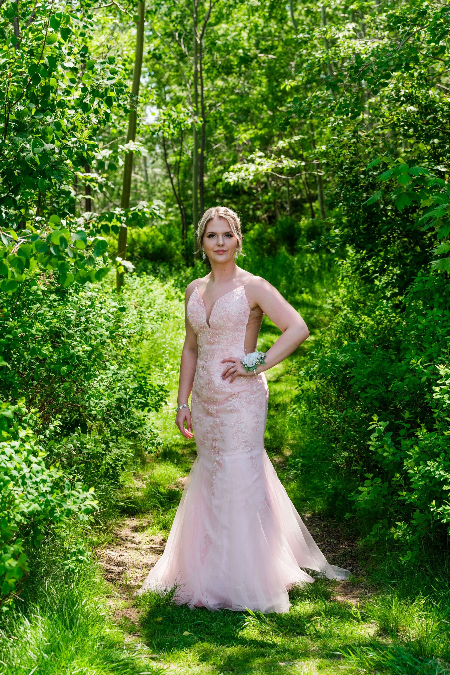 A blush pink mermaid-style gown worn in a lush green forest setting along a natural path.