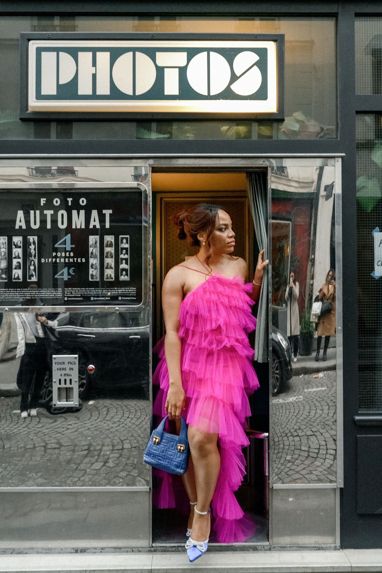 Person in a vibrant pink ruffled dress and blue handbag poses in front of a retro photo booth.