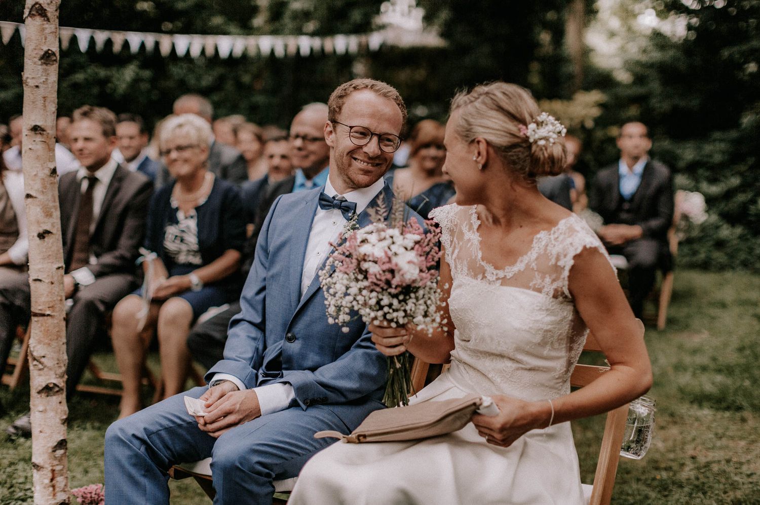 Outdoor garden wedding ceremony with couple seated among bunting decorations.