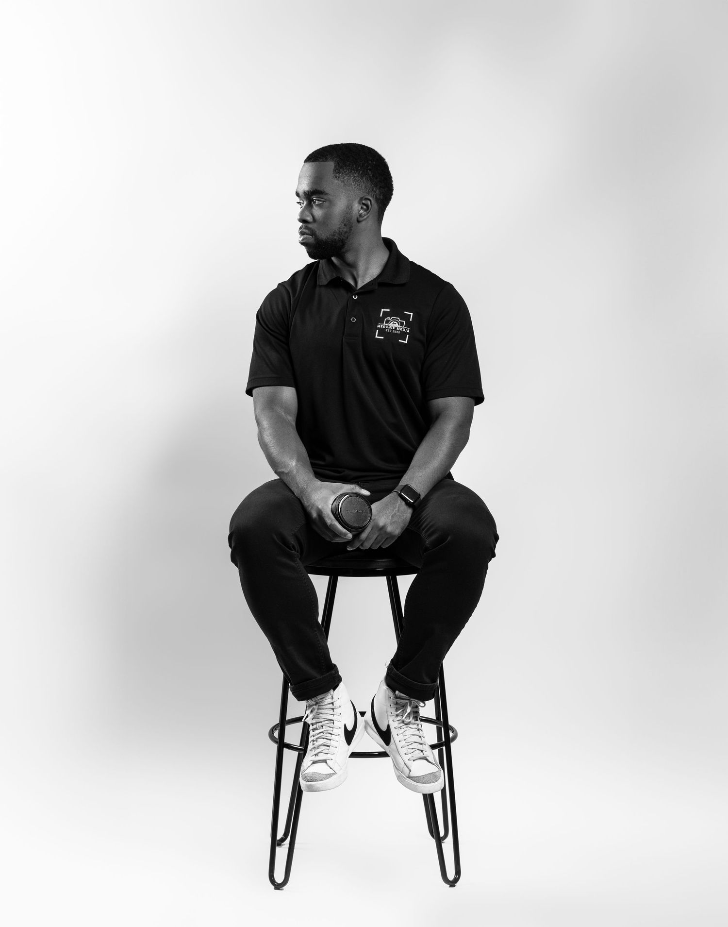 Black and white portrait of person in polo shirt sitting on tall stool against plain background.