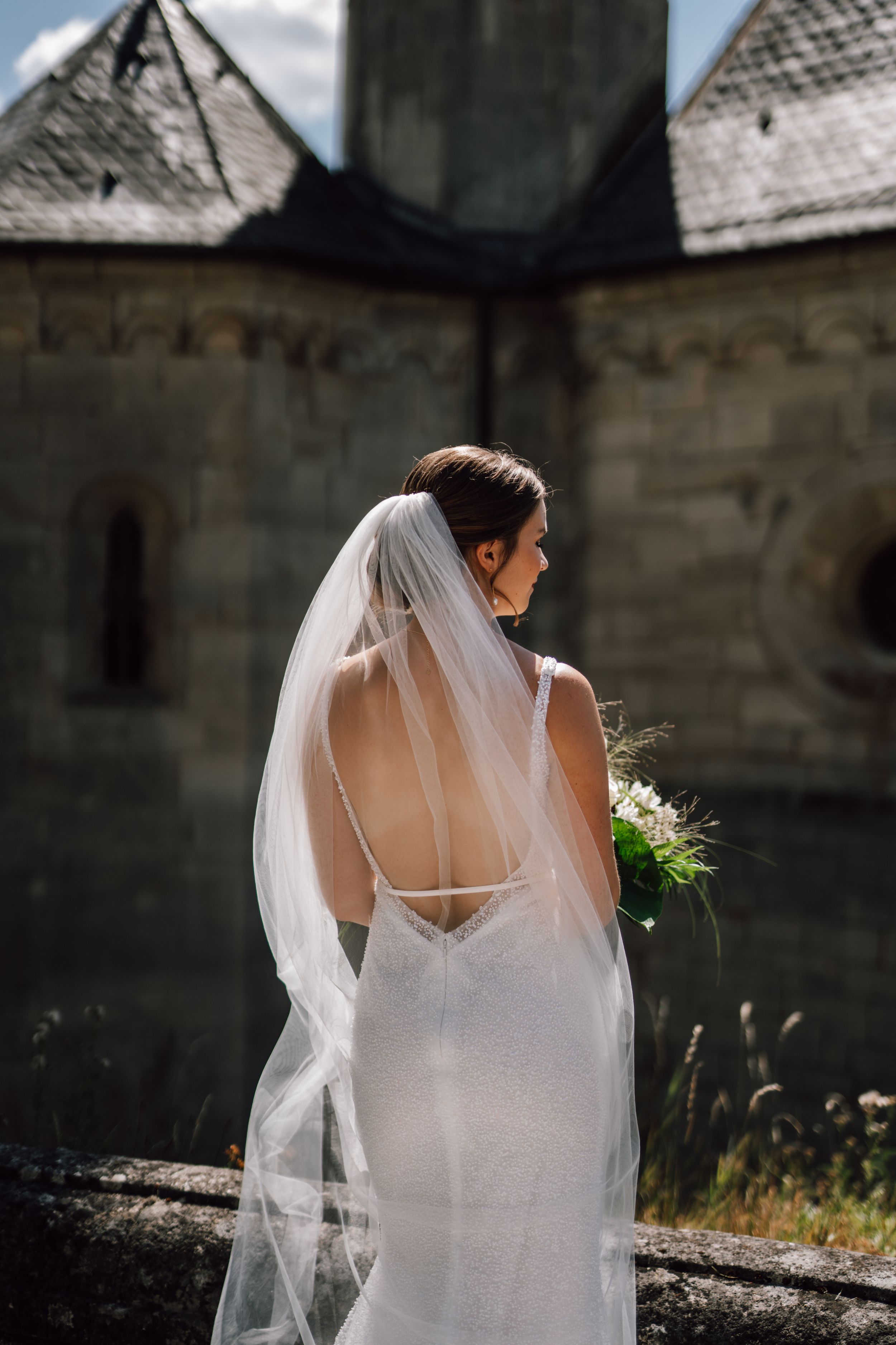 Elegant bridal silhouette in white dress and veil against stone architecture backdrop.