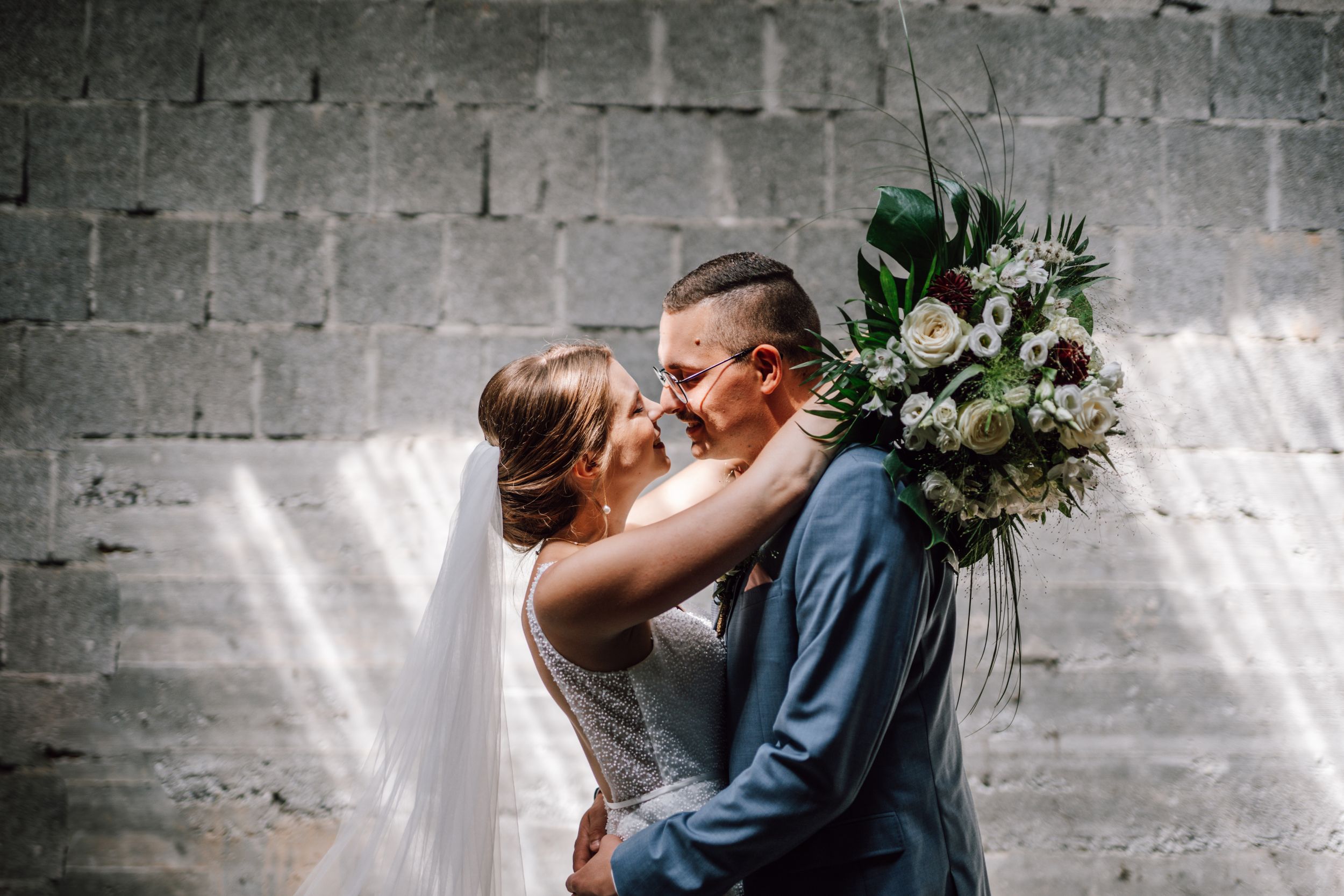 Newlyweds share romantic moment against industrial stone wall backdrop.