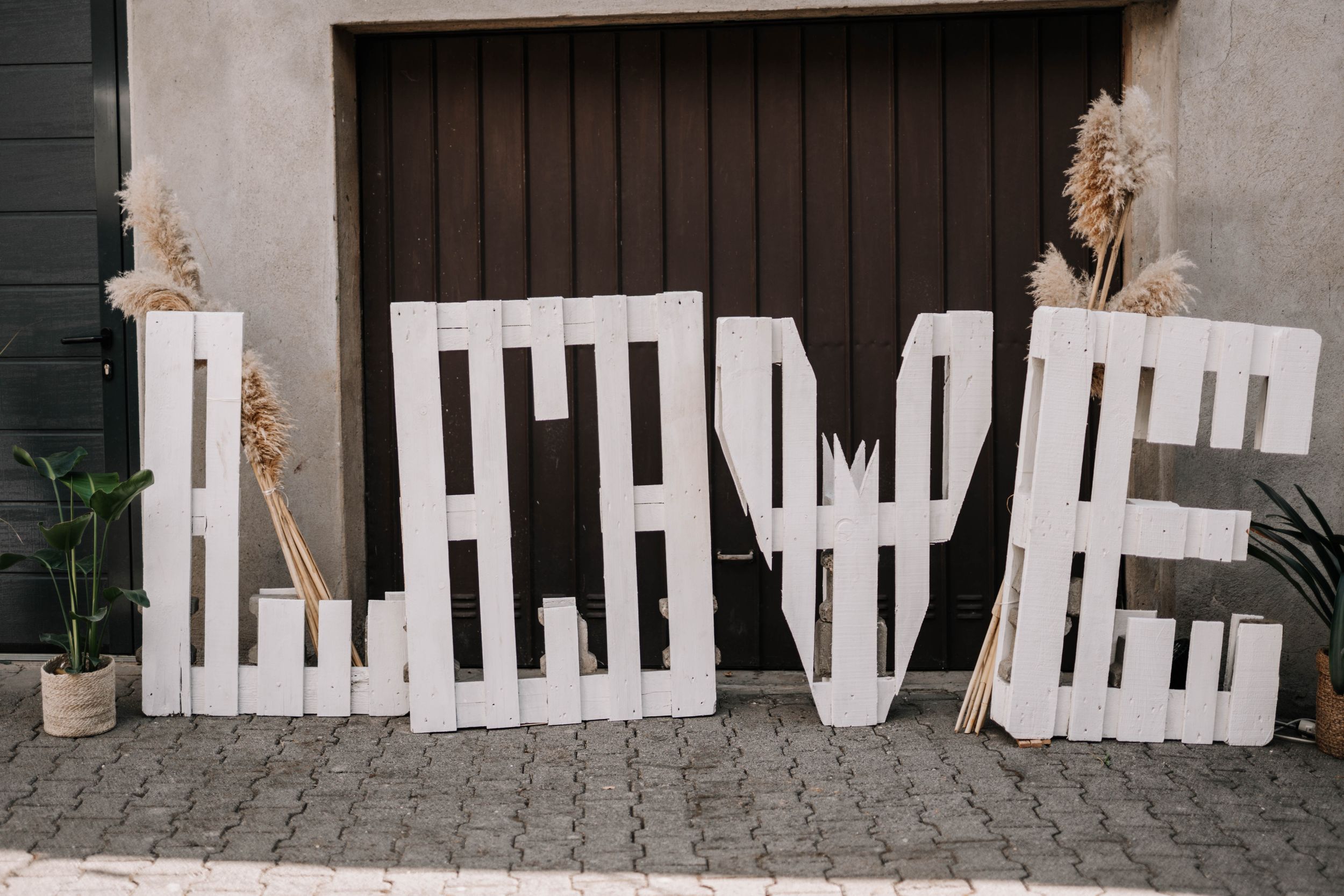 Large white decorative 'LOVE' letters arranged against a dark door with dried pampas grass accents.