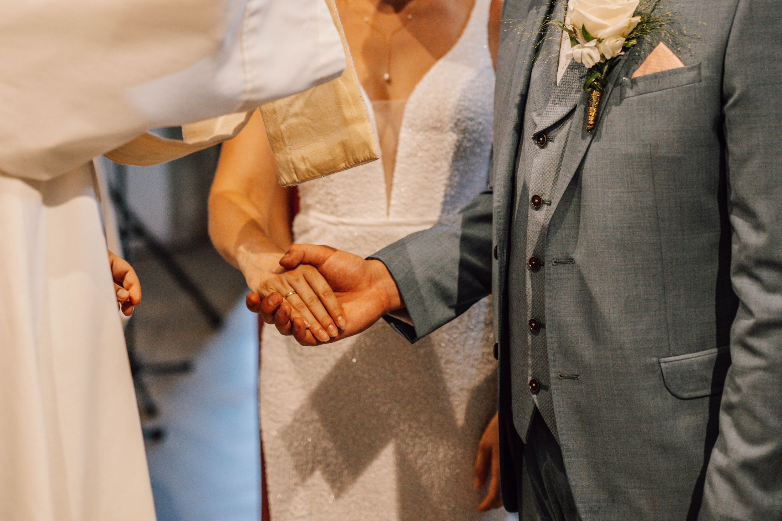 Close-up shot of clasped hands during a wedding ceremony with a grey suit visible.