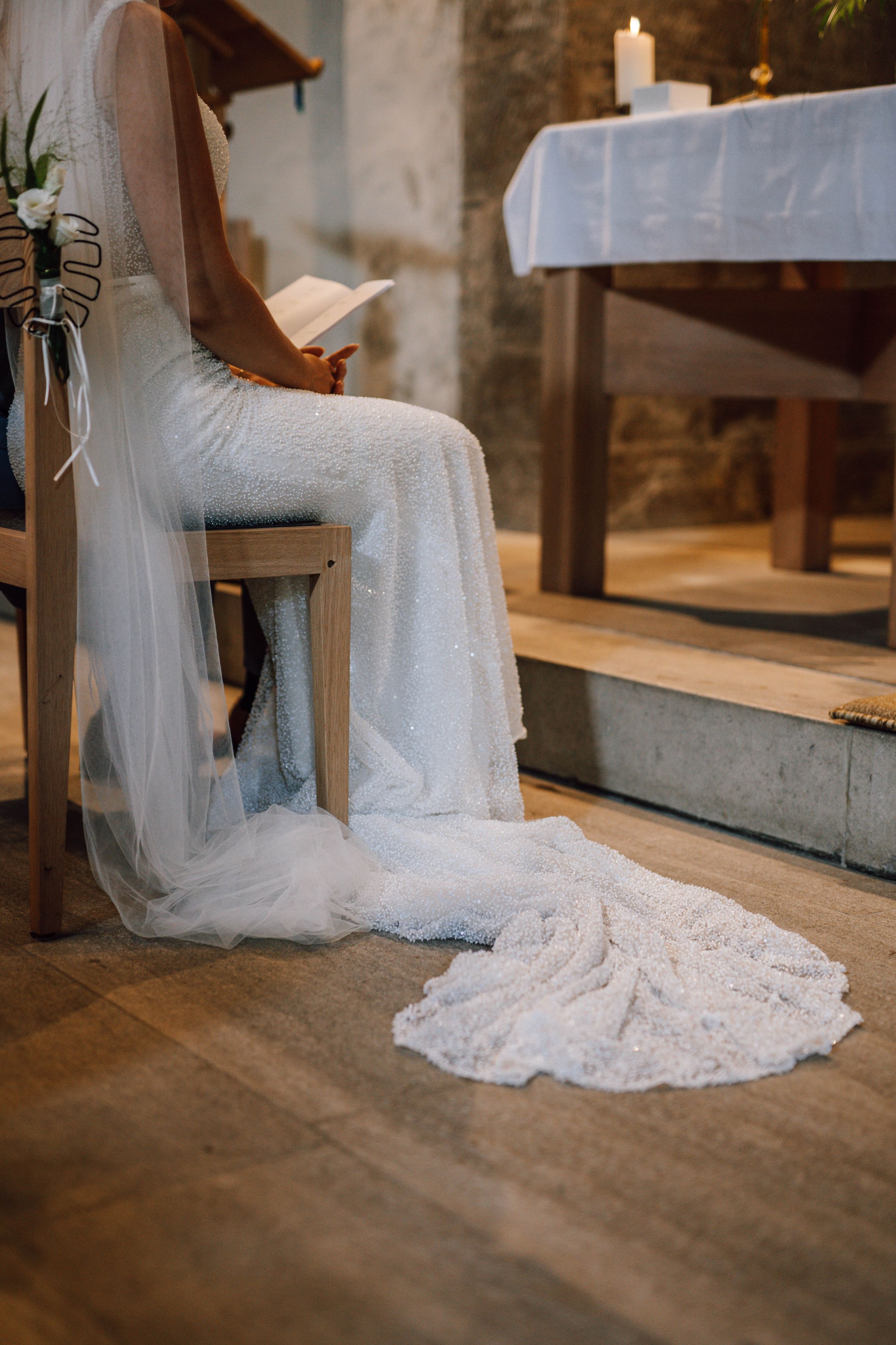 Bride in lace wedding dress sitting with flowing train on wooden church floor.