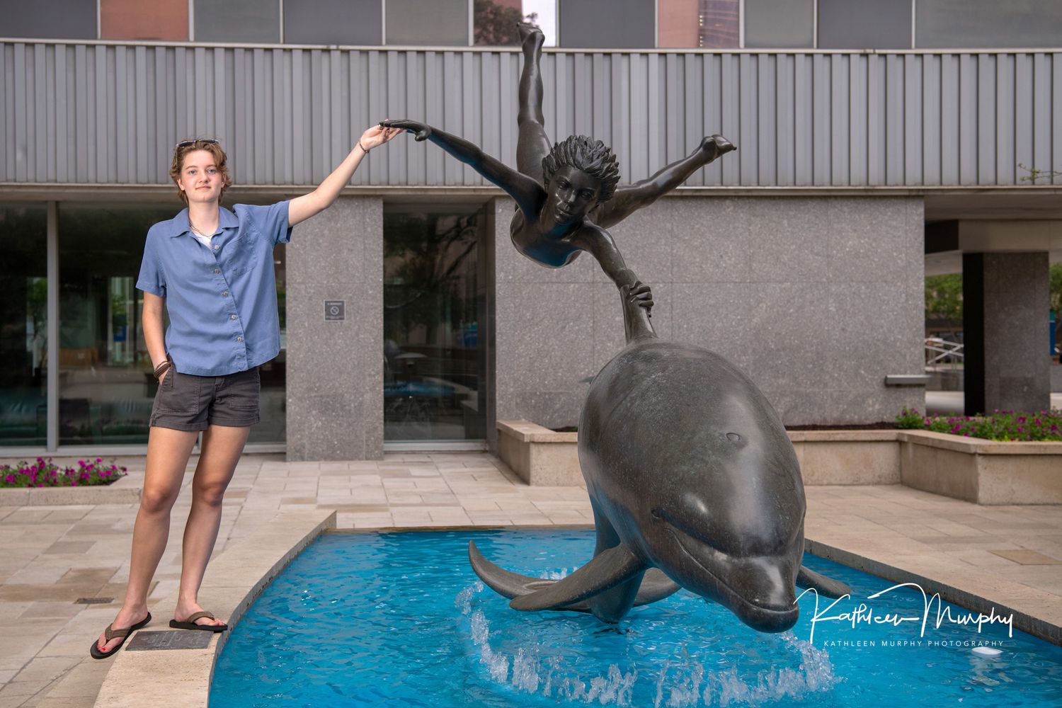 Figure poses next to bronze dolphin fountain sculpture outside office building.