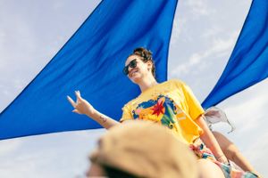 Person in yellow shirt against blue sail background making peace sign.