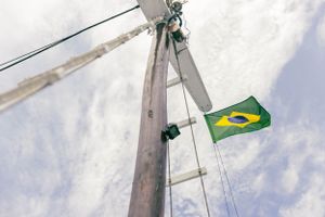 Brazilian flag flying from white sailboat mast against cloudy sky.