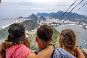 Three friends enjoying scenic view of Rio de Janeiro from cable car overlooking mountains and beaches.
