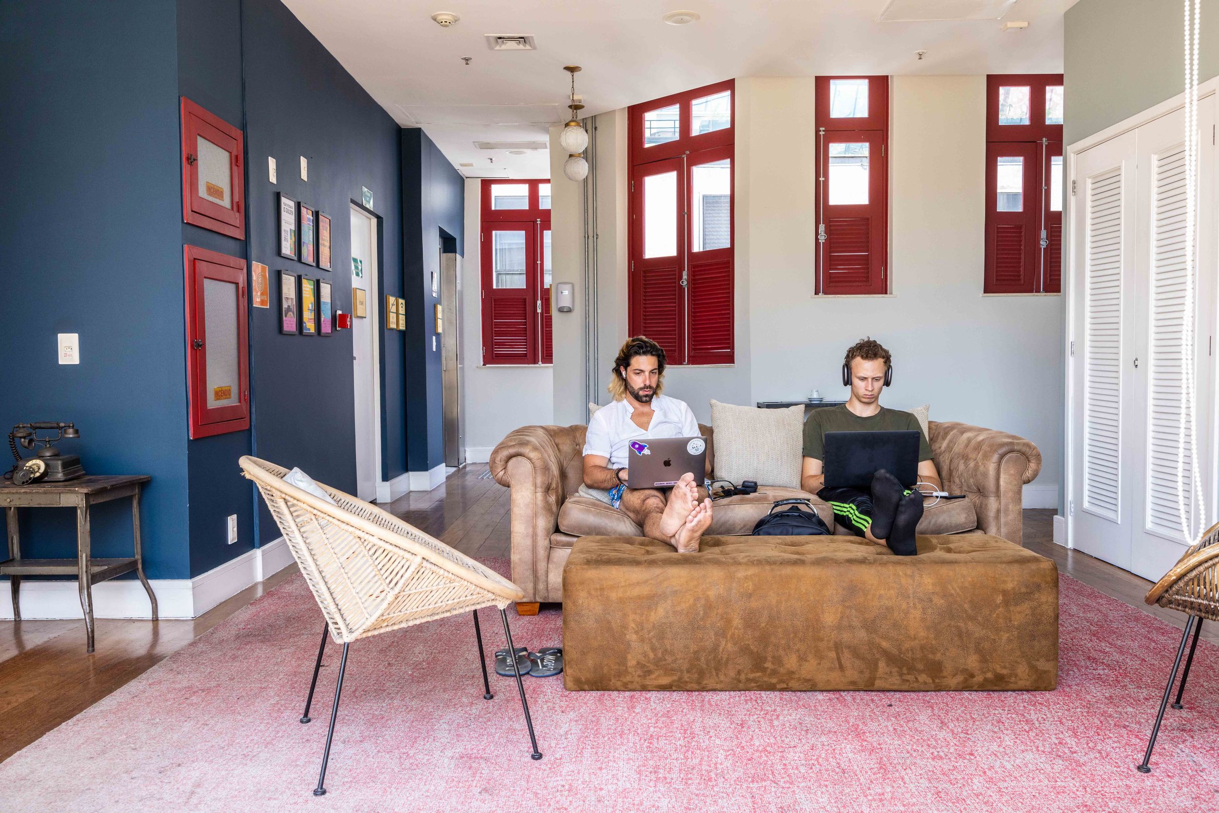 Two people relaxing on a leather couch in a modern hostel lounge with red windows.