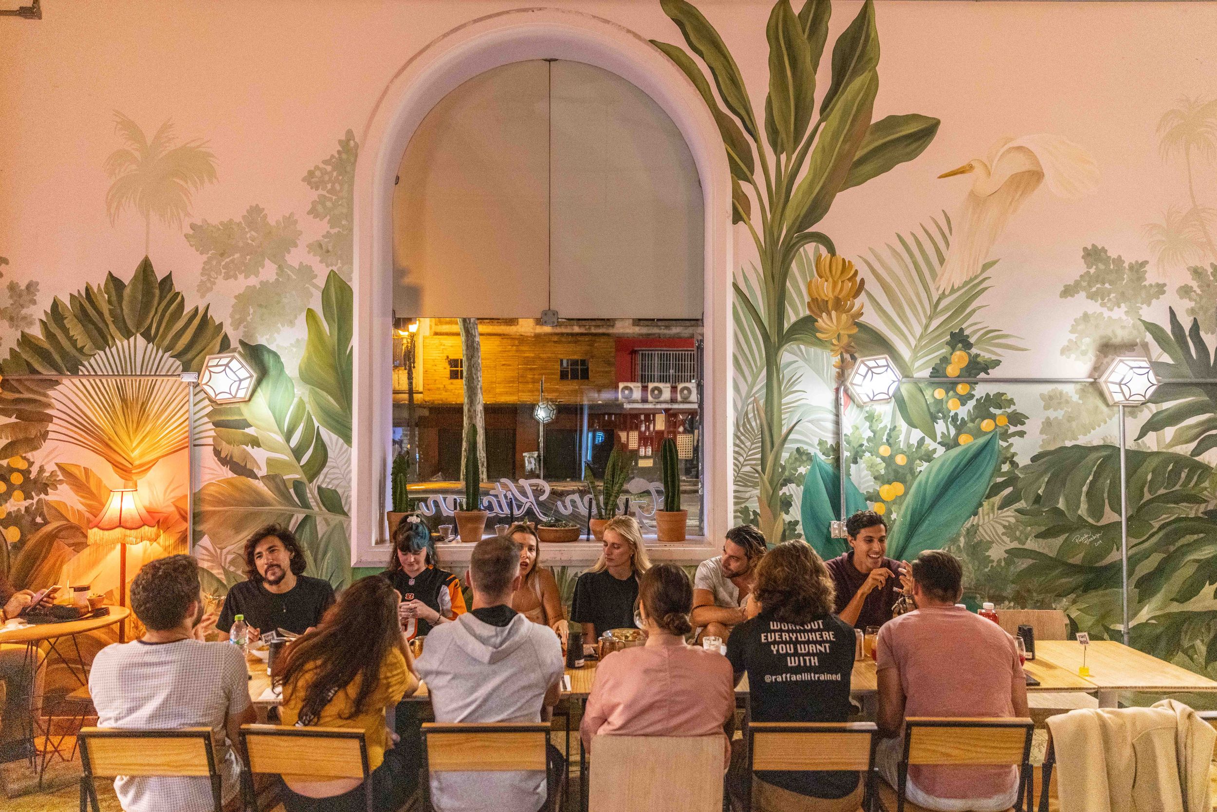 Restaurant interior with tropical wall murals and diners seated beneath an arched doorway.