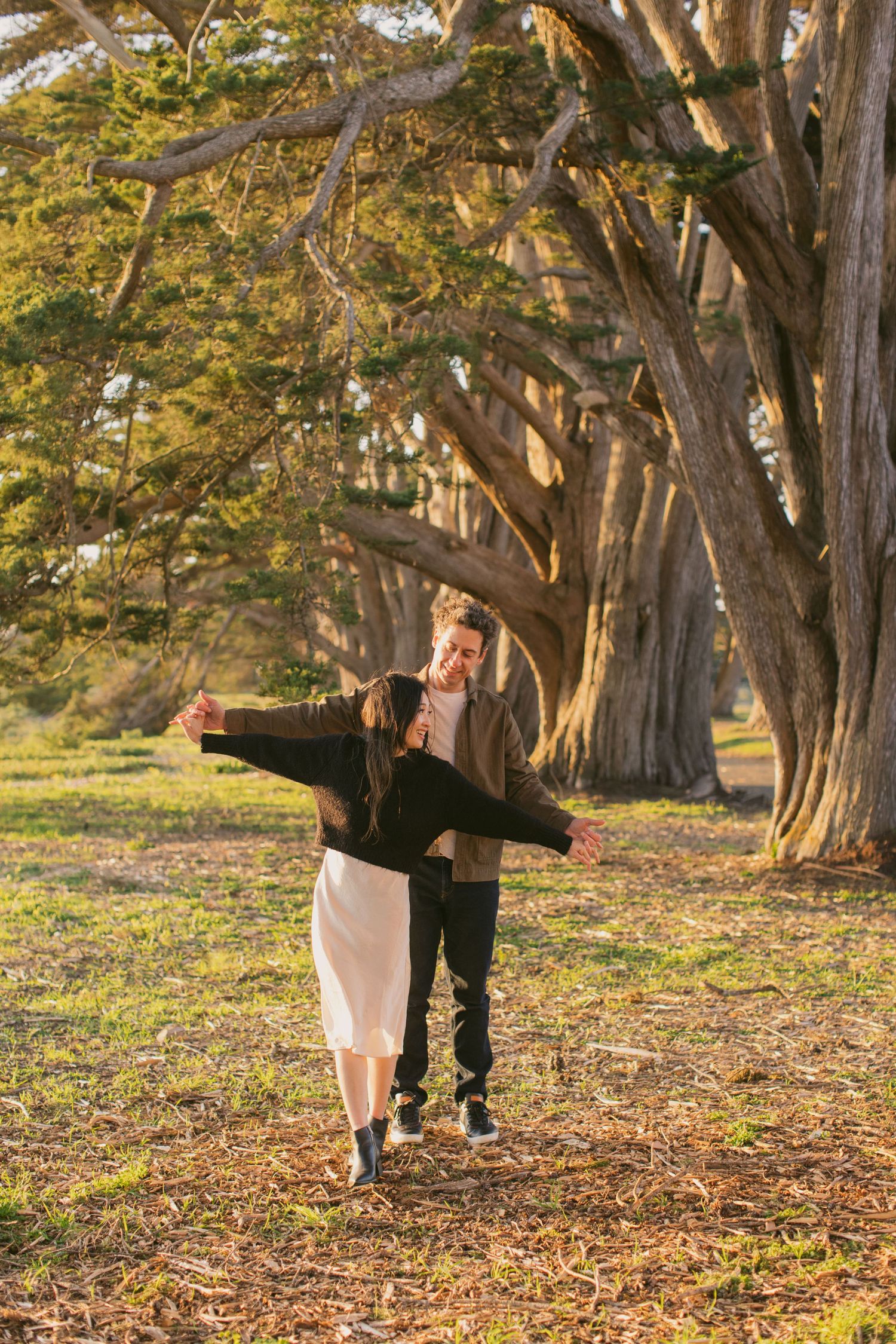 A couple playfully spreads their arms while walking through a tree-lined path.