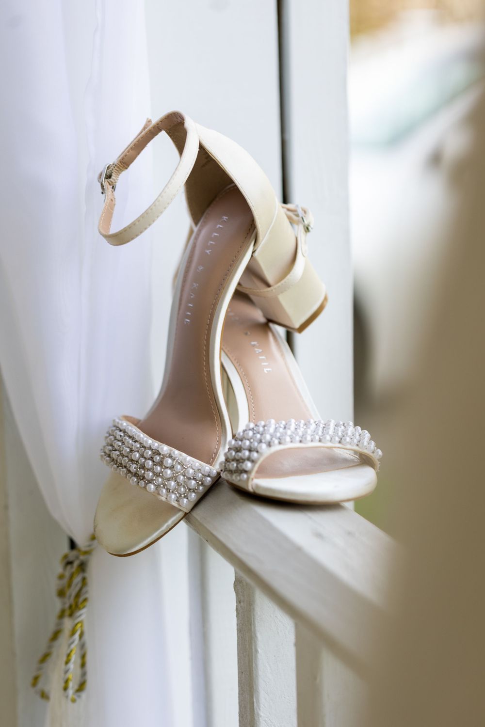 Elegant white bridal shoes with crystal embellishments displayed on windowsill.