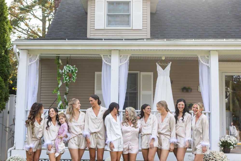 Group of bridesmaids in matching white robes sitting on porch with wedding dress hanging.