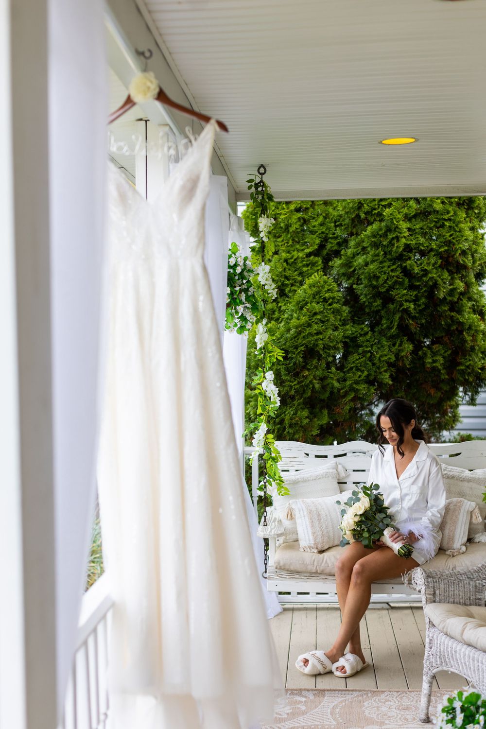 Wedding dress hanging on porch with person relaxing on white wicker furniture.