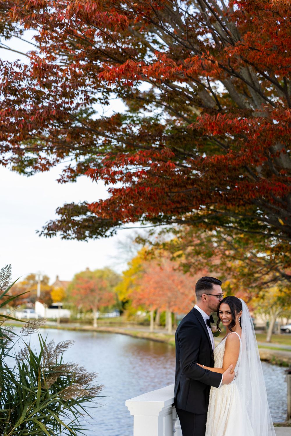 A couple embraces beneath vibrant red autumn foliage beside a waterfront path during their wedding photoshoot.