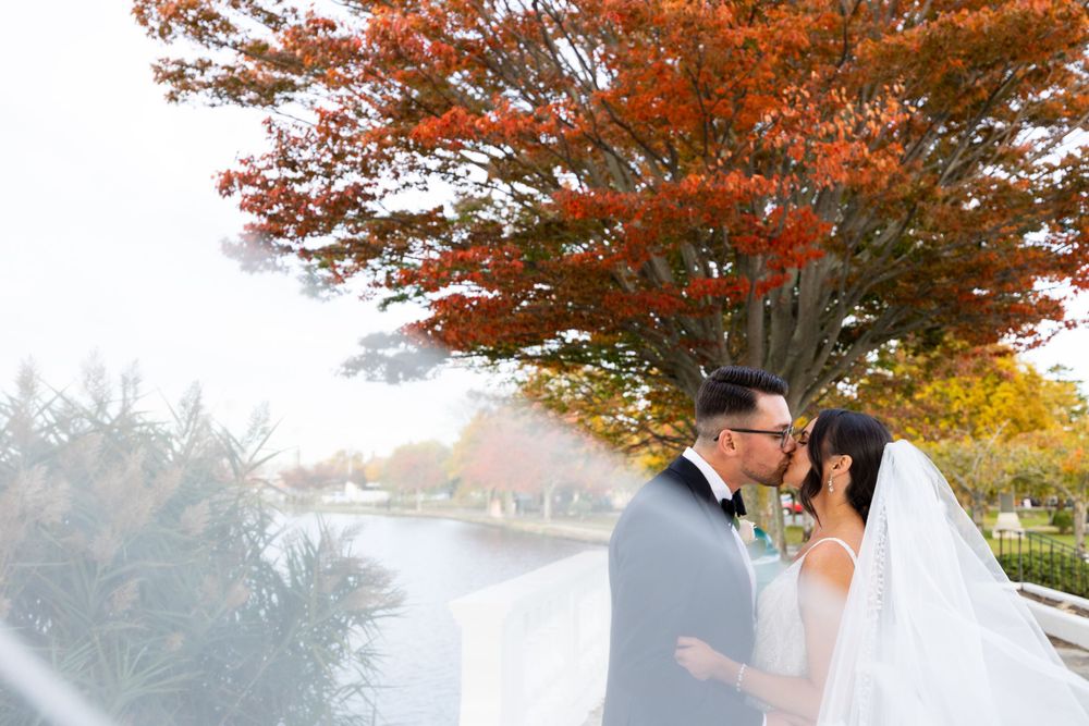 Newlyweds pose along waterfront walkway with dramatic fall foliage overhead.