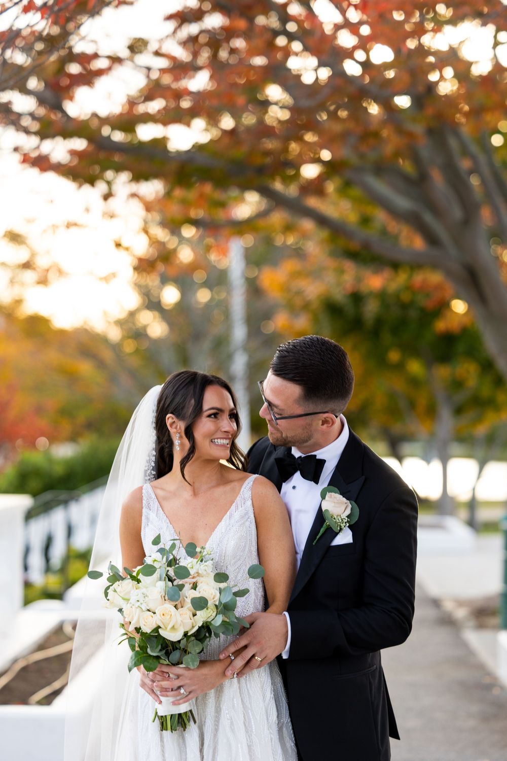Bride holding white bouquet smiles while sharing romantic moment with groom under autumn trees.