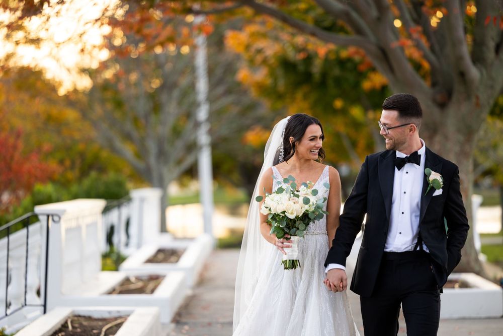 Wedding couple walks hand in hand along white railing pathway with autumn trees in background.
