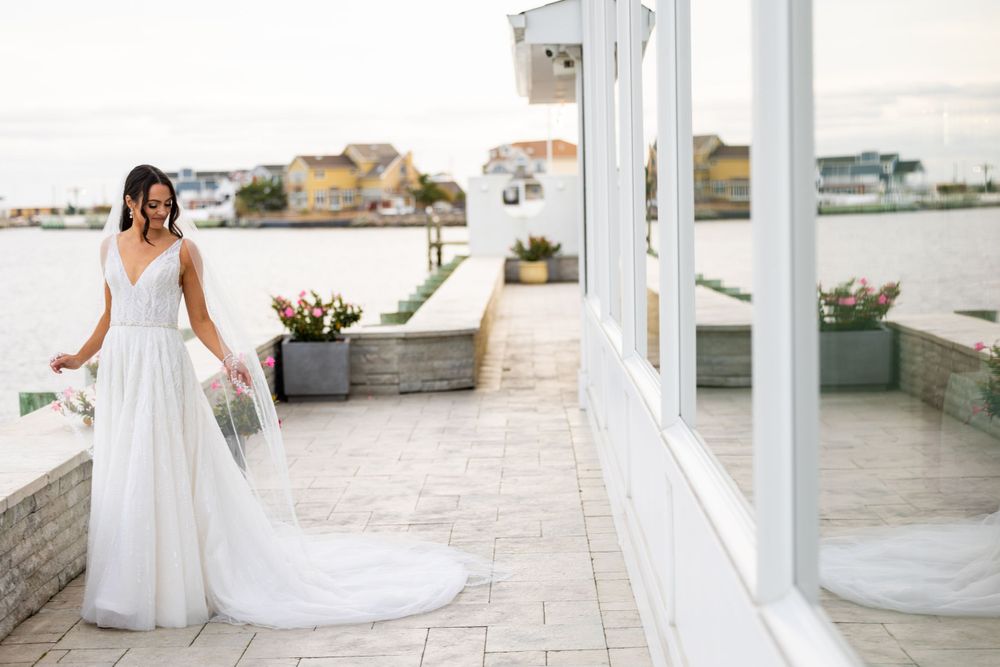 Bride in flowing white dress stands on seaside terrace with trailing veil.