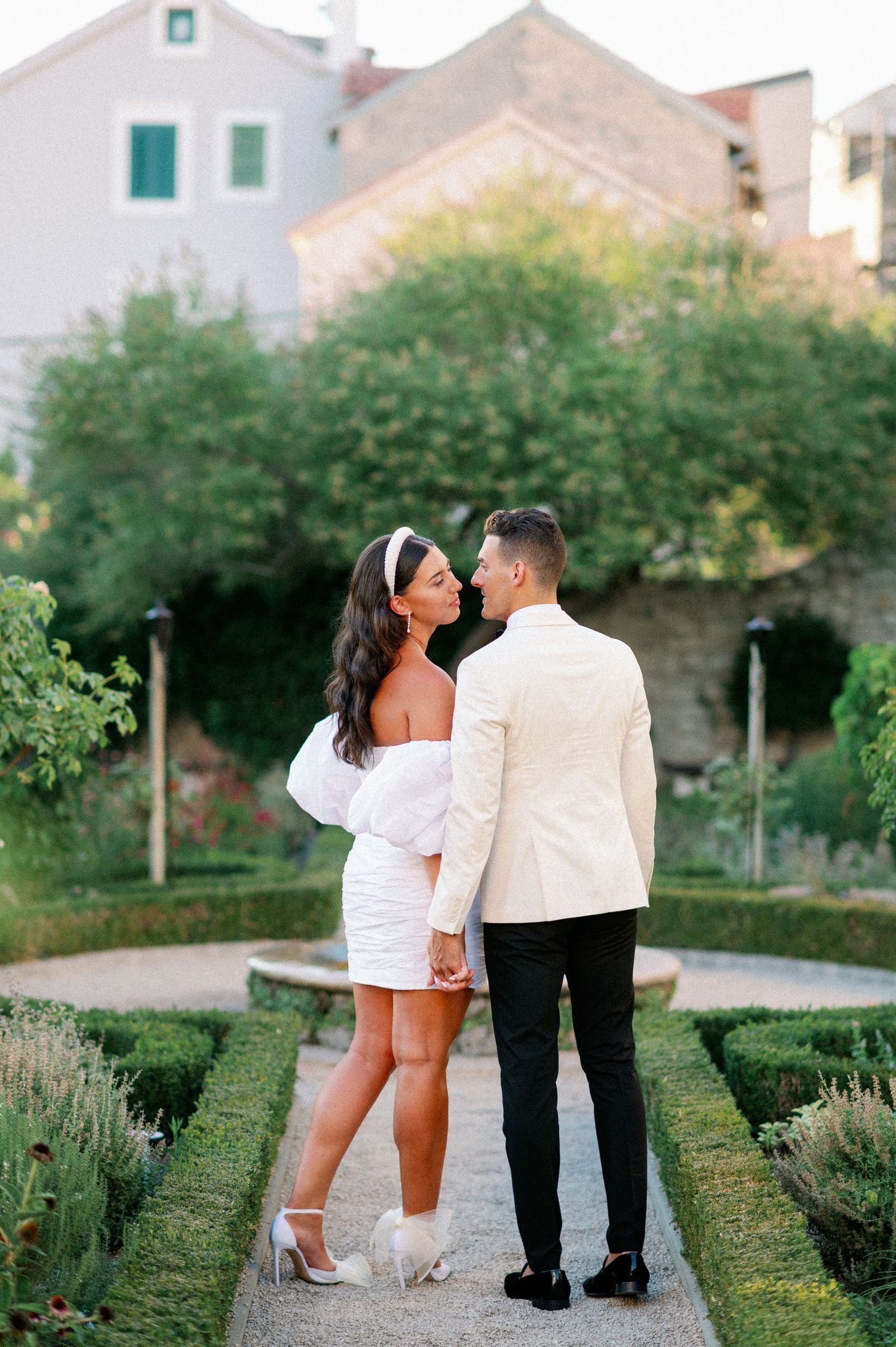 A couple in elegant white formal wear share a romantic moment in a lush garden path.