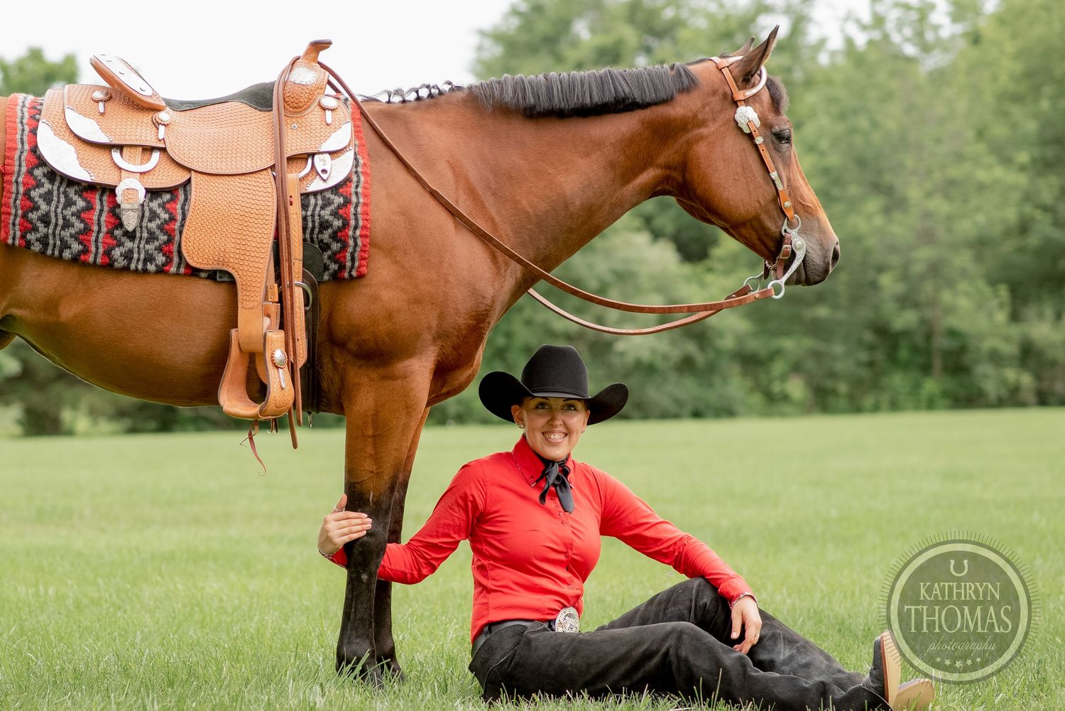 Western-saddled brown horse stands while handler sits in grassy field.