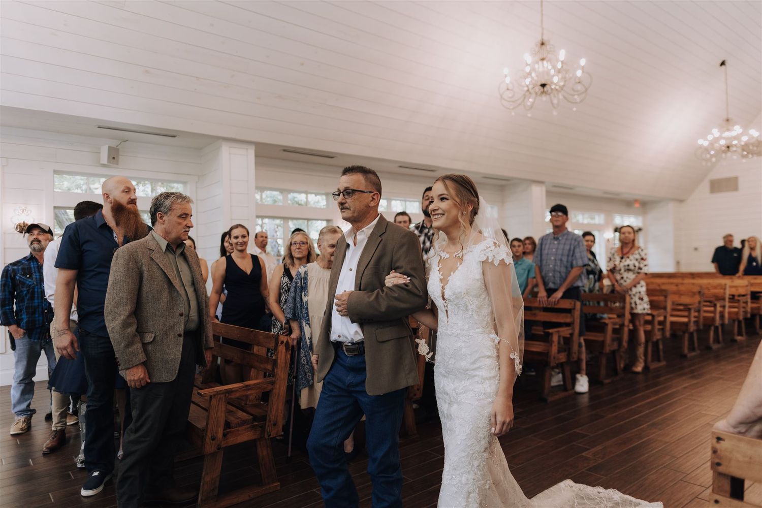 Wedding ceremony in bright chapel with wooden pews and chandeliers.