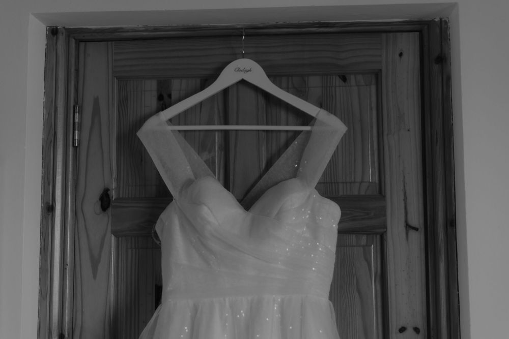 A white wedding dress hangs elegantly on a wooden hanger against a dark doorframe.