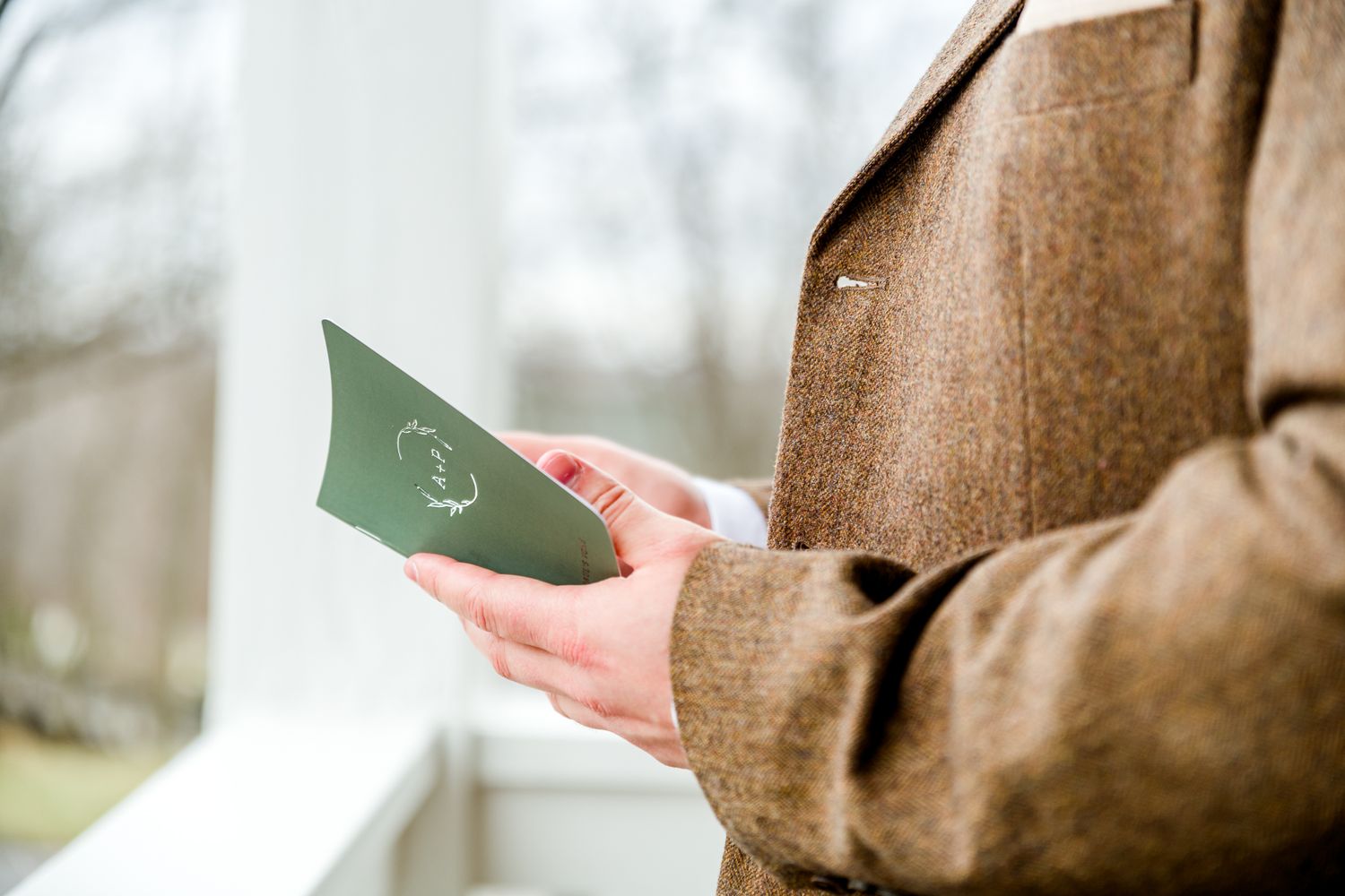 A man in a brown suit holds a small green vow book outdoors by Stephanie Coulter Photography