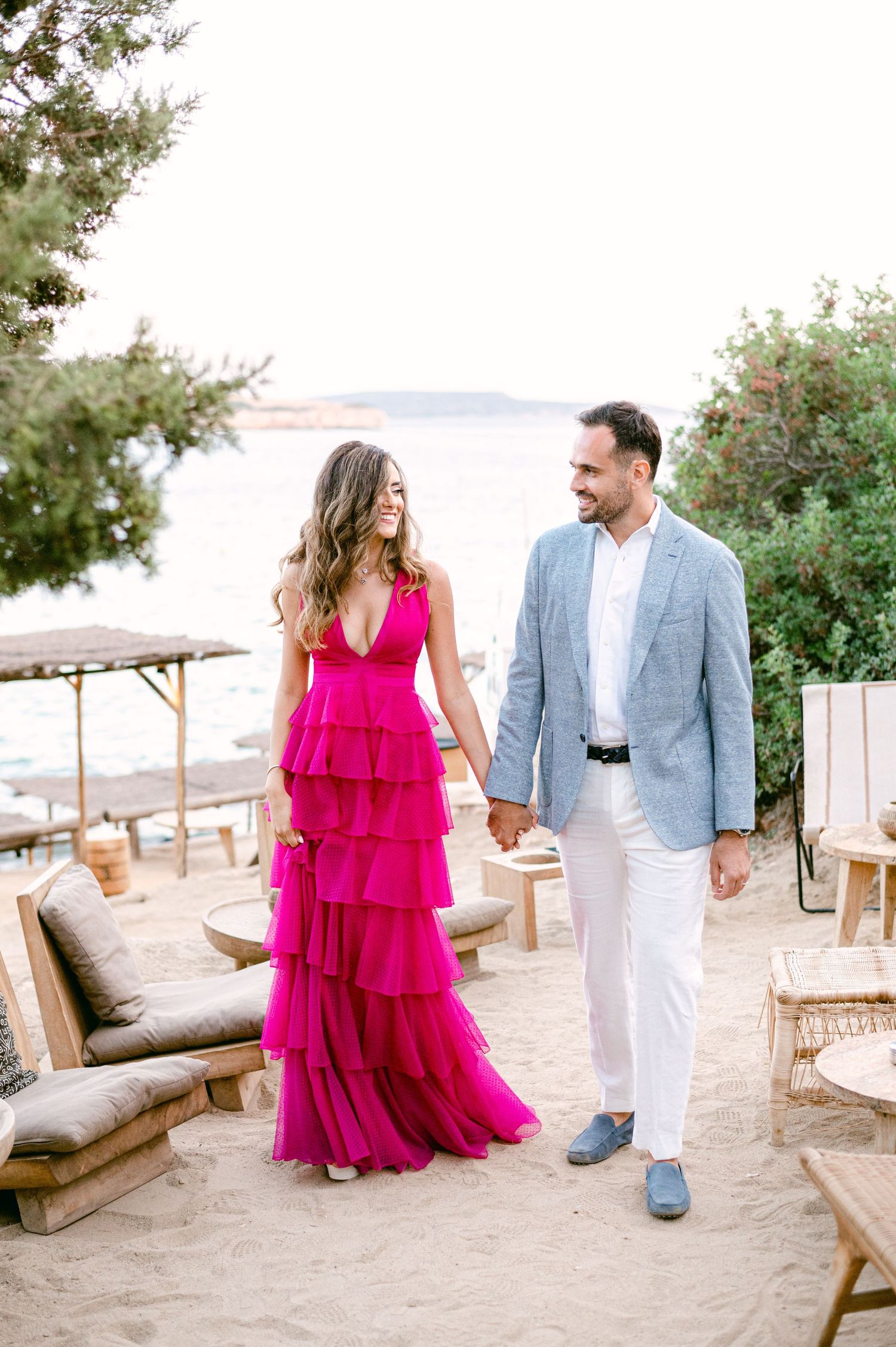 Couple walking hand in hand on beach path with rustic outdoor seating and ocean view.