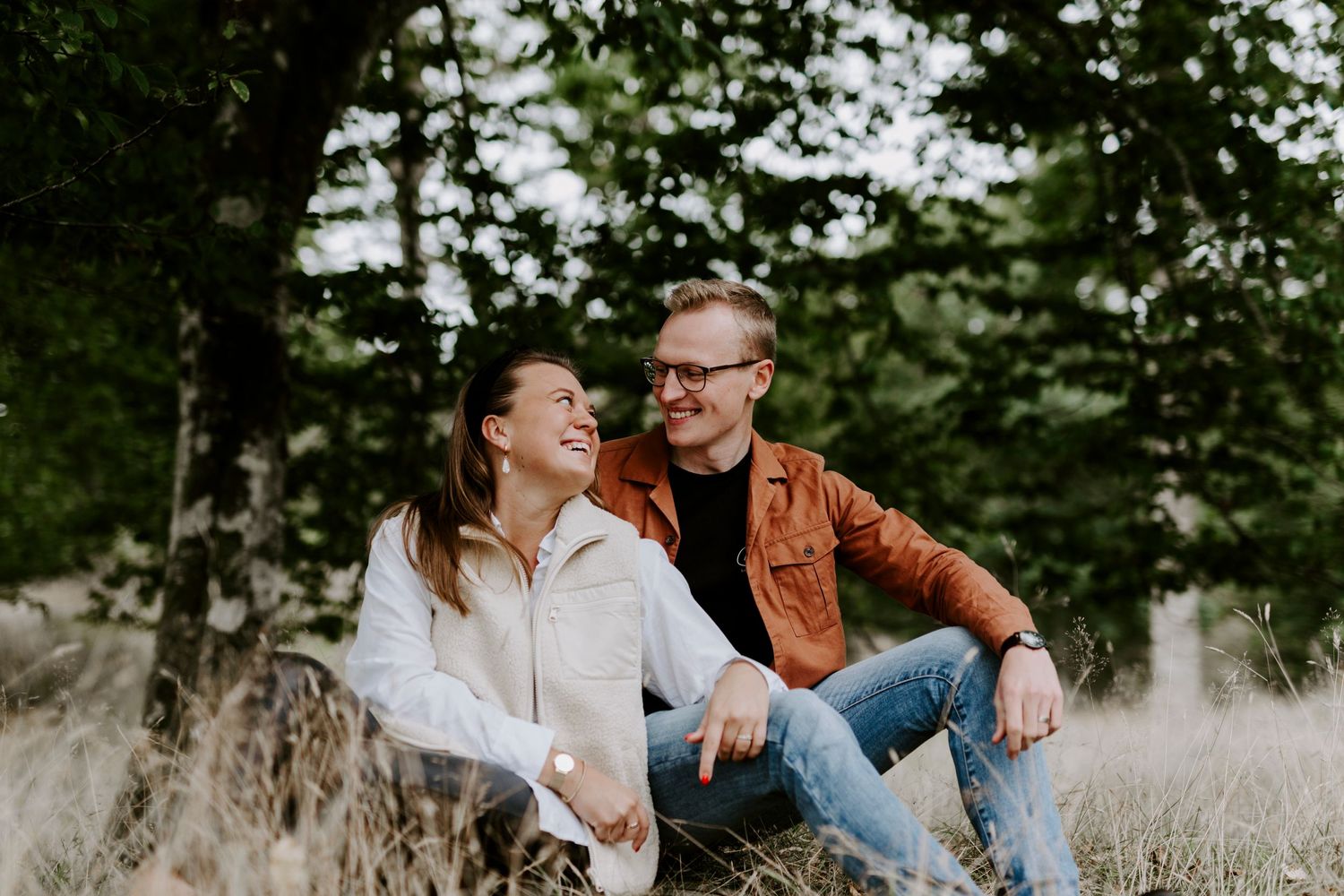 Candid couple sitting in tall grass with trees in background during outdoor photoshoot.