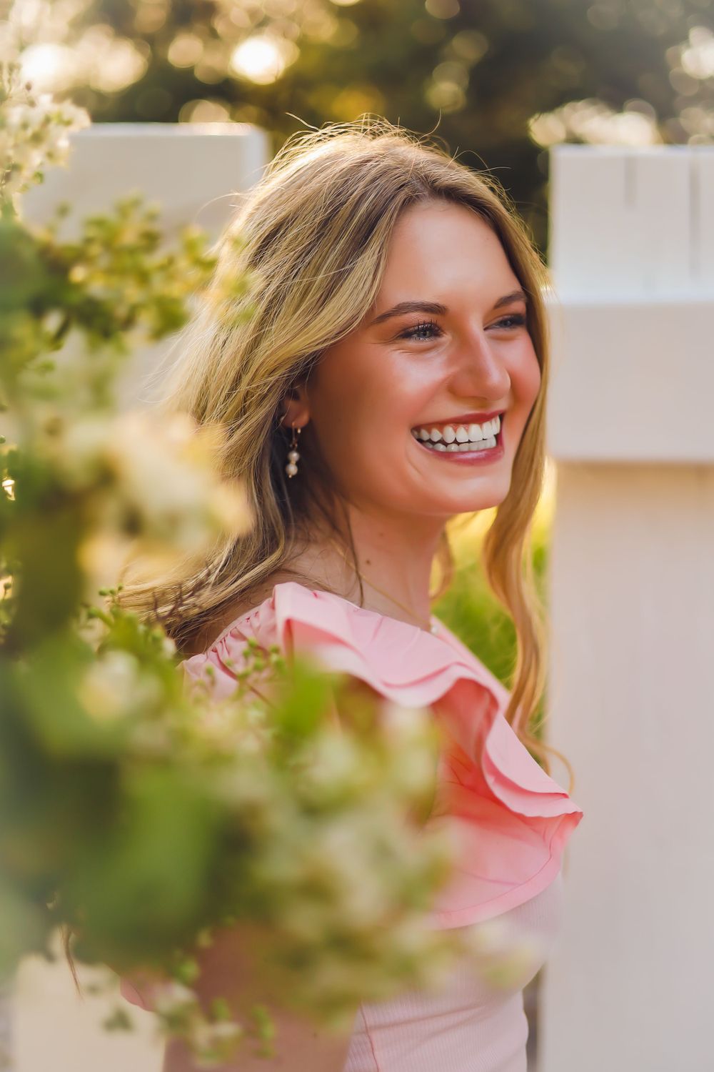 Sun-lit portrait of woman in pink ruffled dress smiling through foliage.