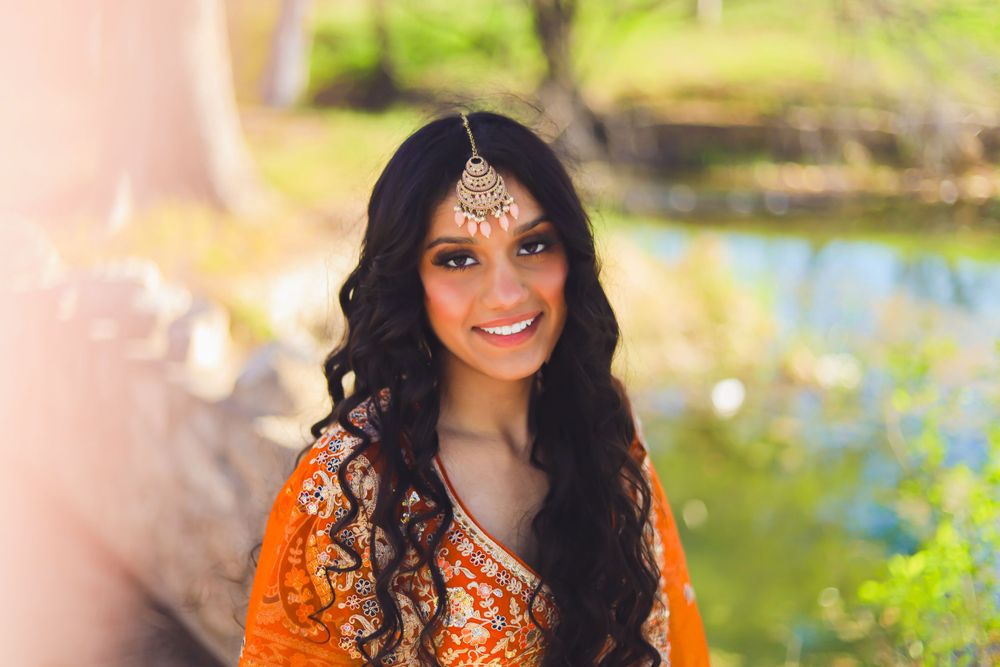 Woman in ornate orange traditional Indian attire and jewelry poses outdoors near water during golden hour.