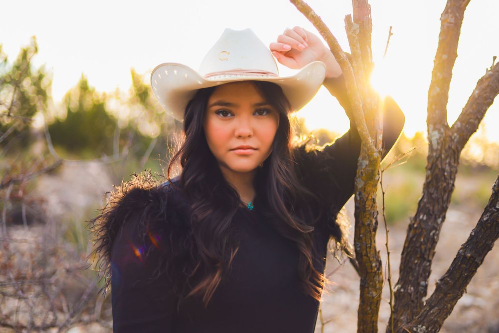 Woman in black sweater and cowboy hat poses against tree during golden sunset.