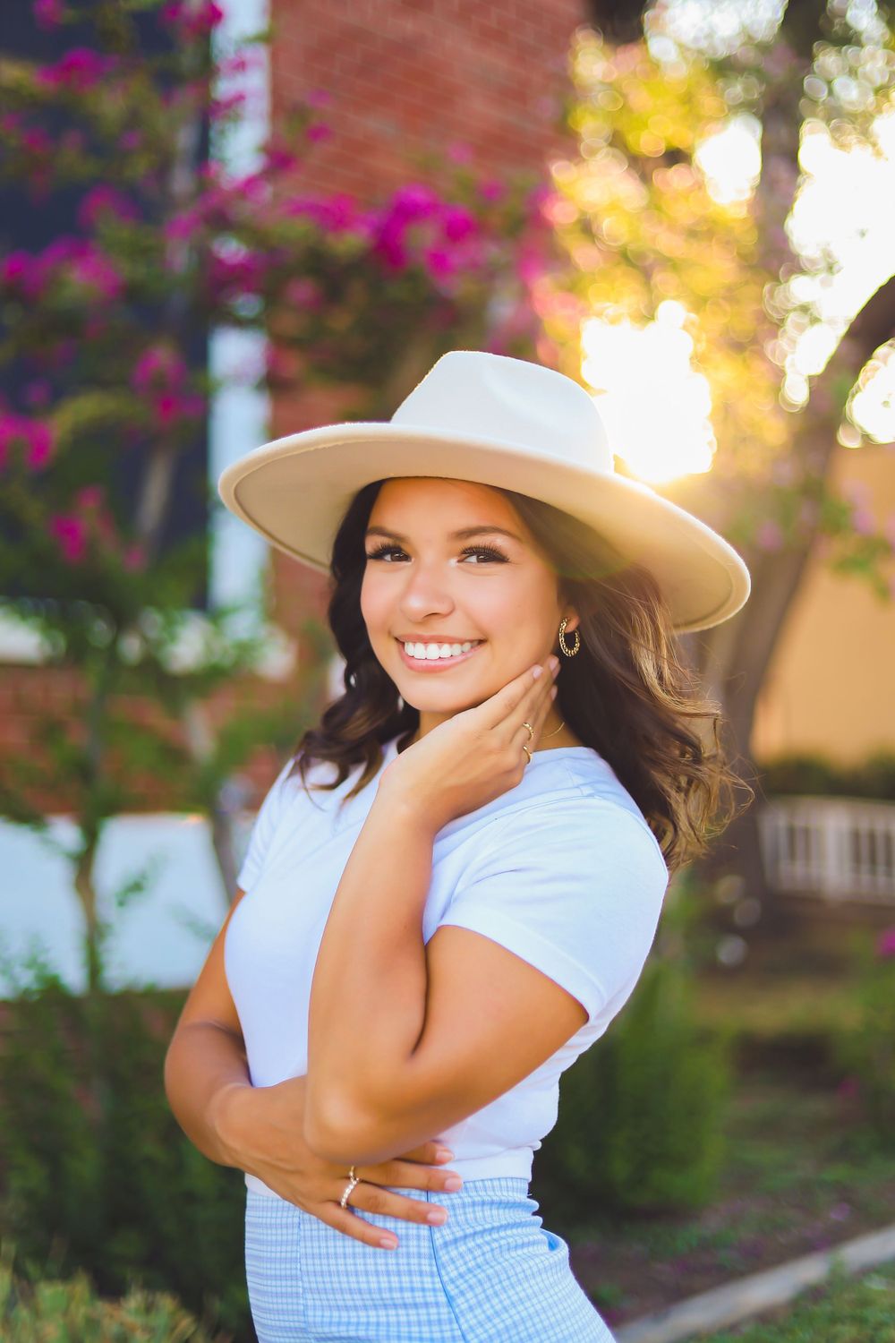 Woman in white hat and gingham skirt poses in garden with blooming flowers.