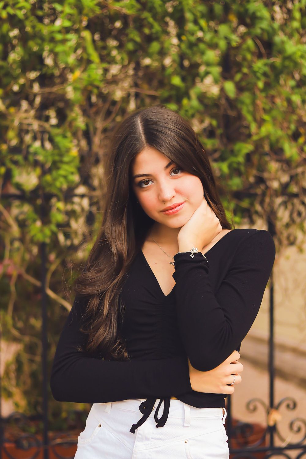 A casual portrait with dark long hair against a leafy background wearing a black top and white pants.