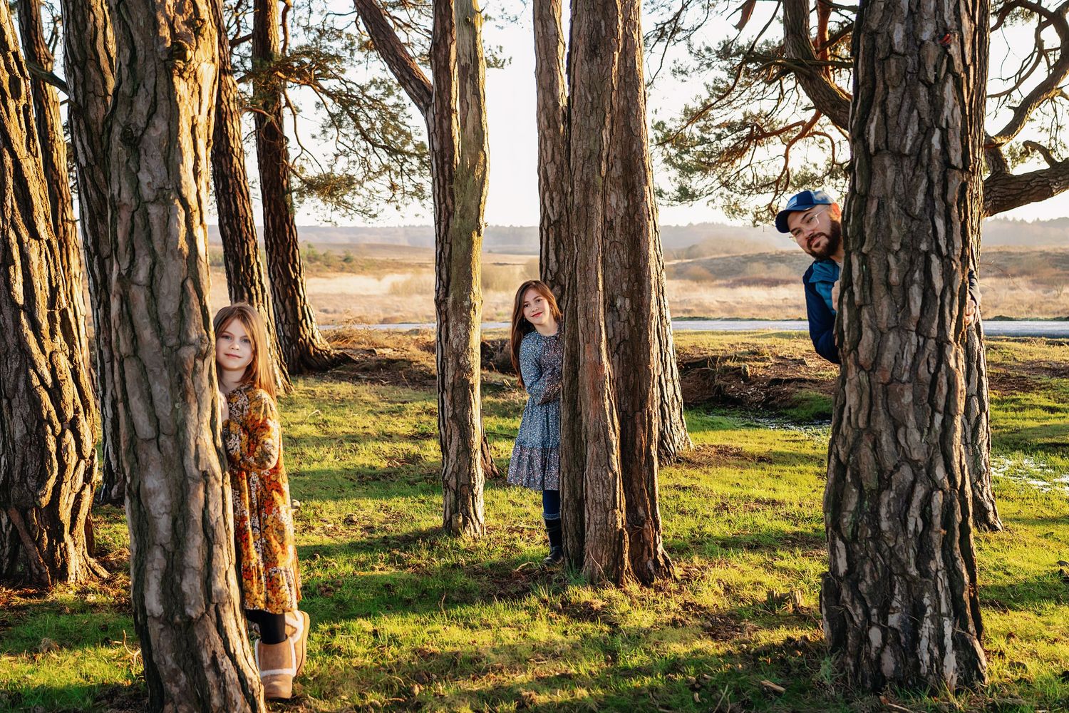 Dad and 2 little girls hiding behind trees laughing and smiling, in the New Forest, Hampshire.