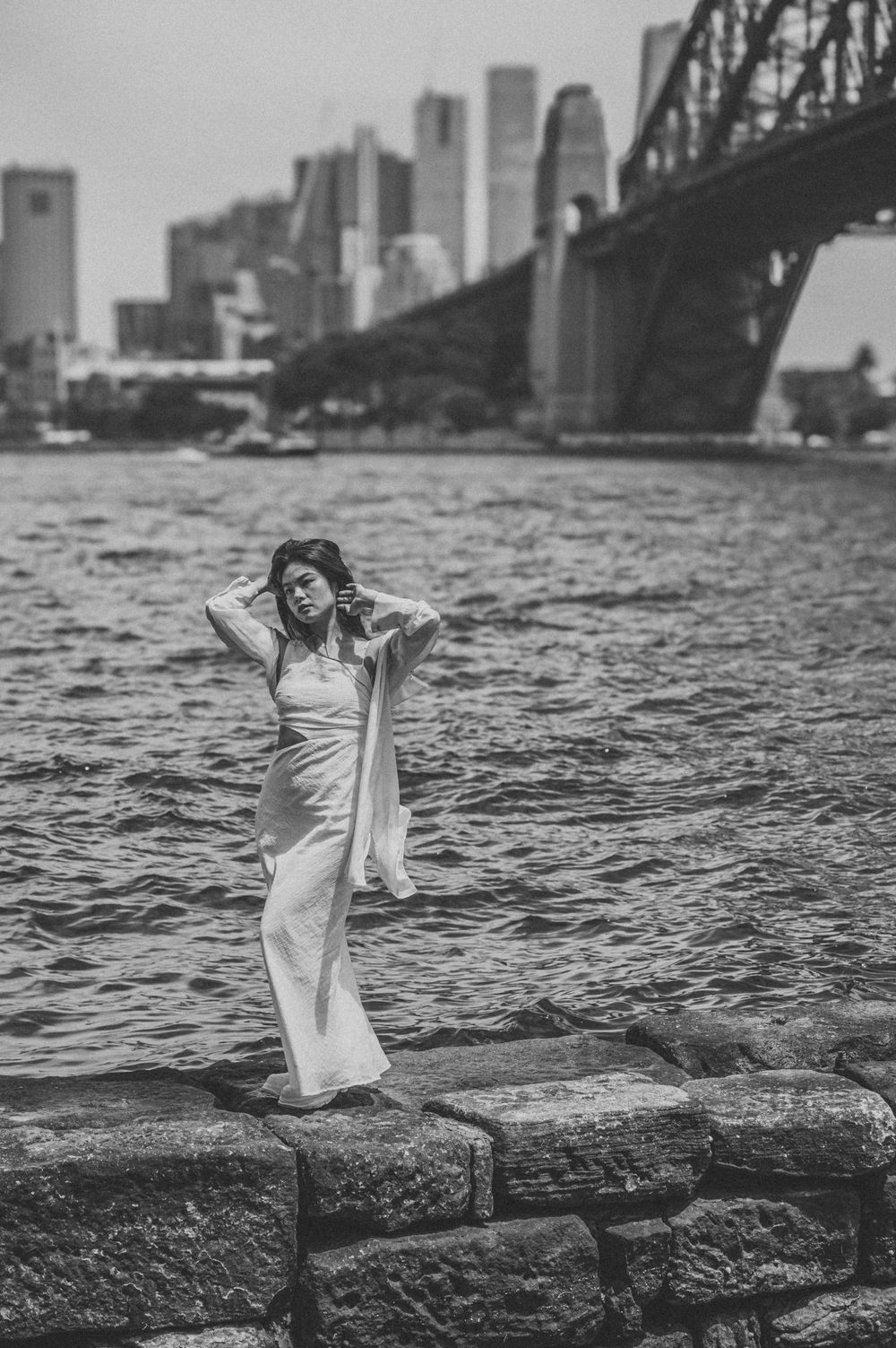 A person in a flowing white dress poses near the Sydney Harbour Bridge in black and white. SIX84 Photography, Portrait Photography, Sydney