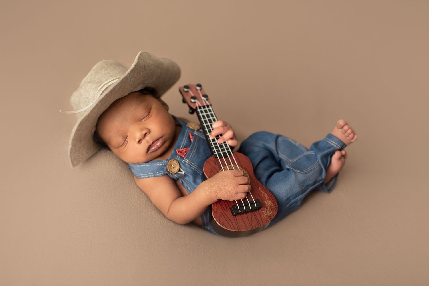 Sleeping newborn wearing denim overalls and cowboy hat while holding tiny guitar on beige backdrop.