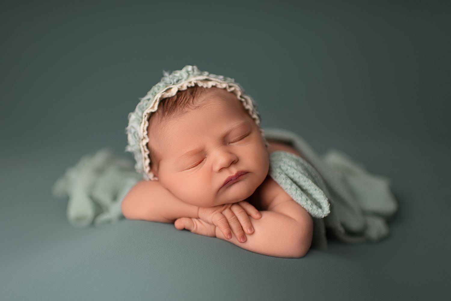 Sleeping newborn wearing lace headband posed on mint green background in classic pose.