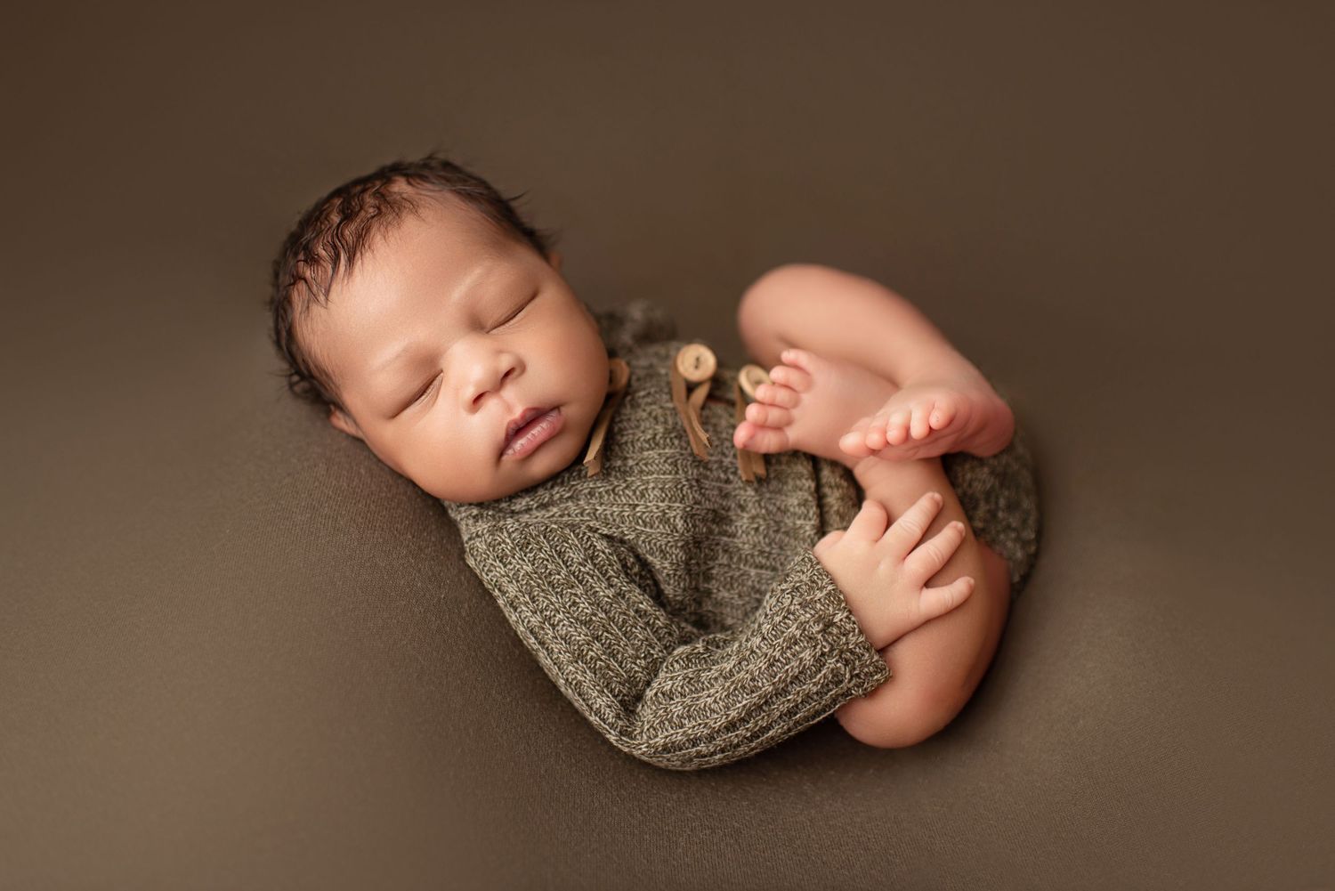 Sleeping newborn wearing gray knit sweater posed on brown background.